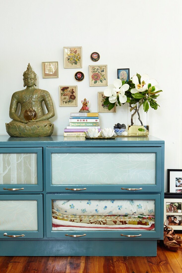 Buddha statue on blue sideboard with glass drop-down doors below framed pictures on wall