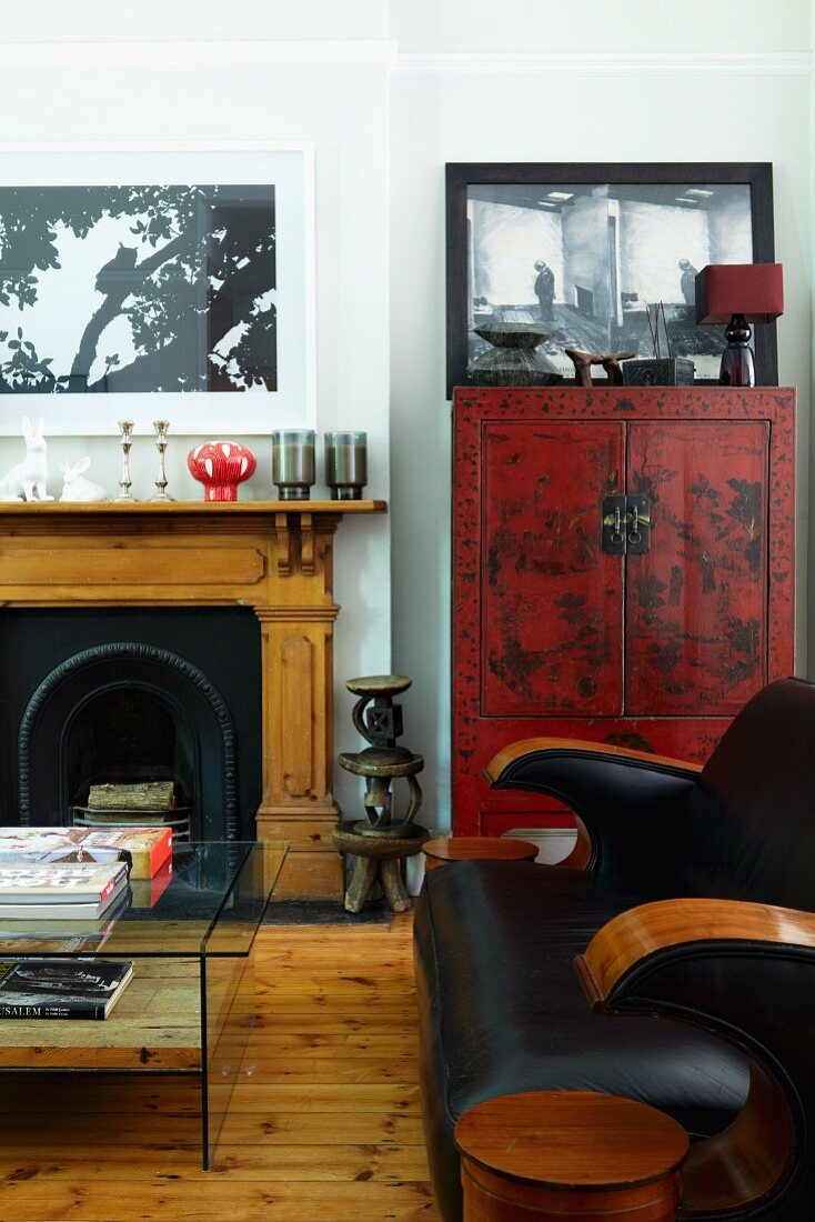 Black leather couch and glass table in front of open fireplace and red-painted, Oriental cabinet in traditional living room