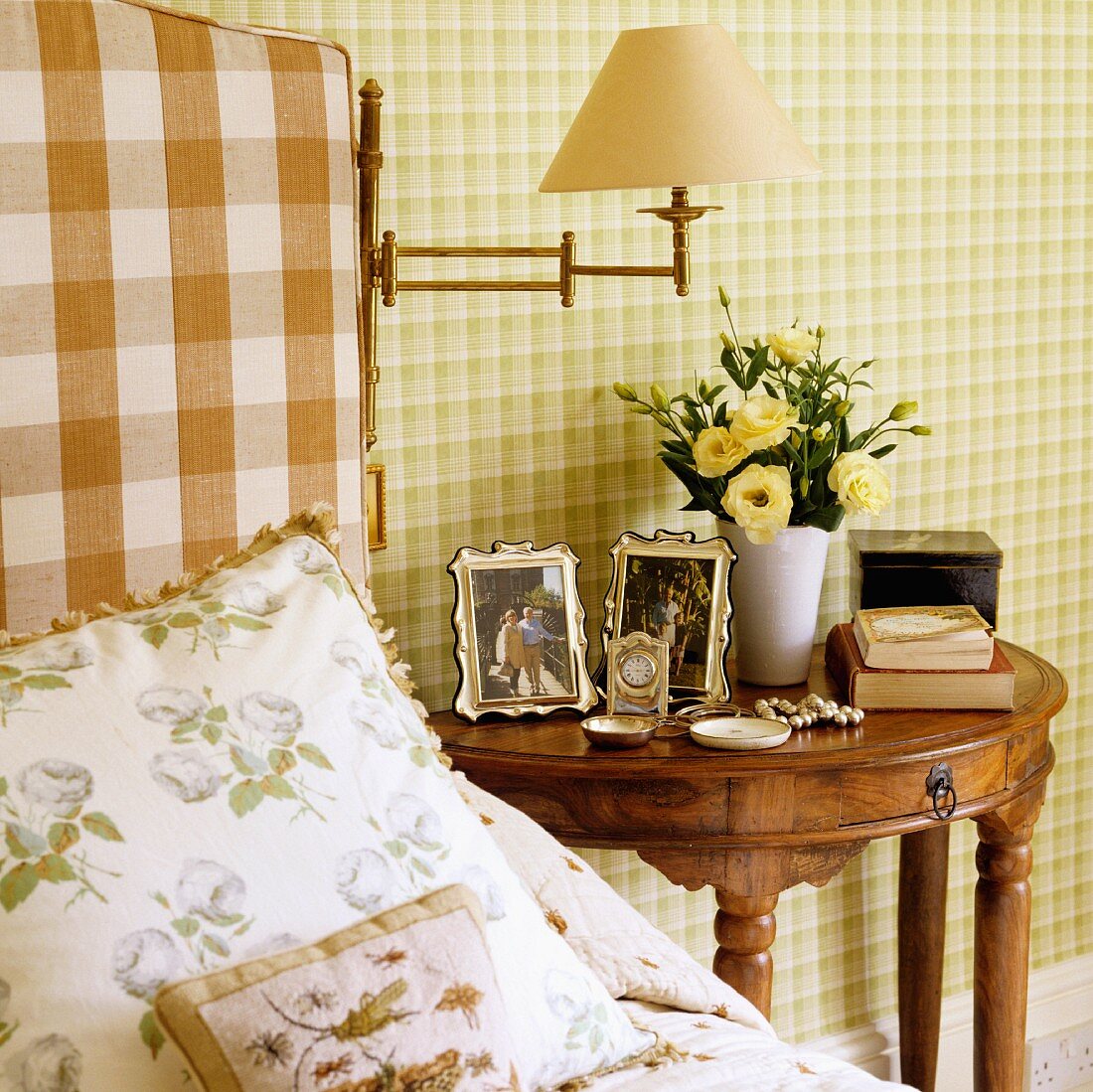 Scatter cushions on bed next to antique, semi-circular bedside table in exotic wood against wall with green checked wallpaper
