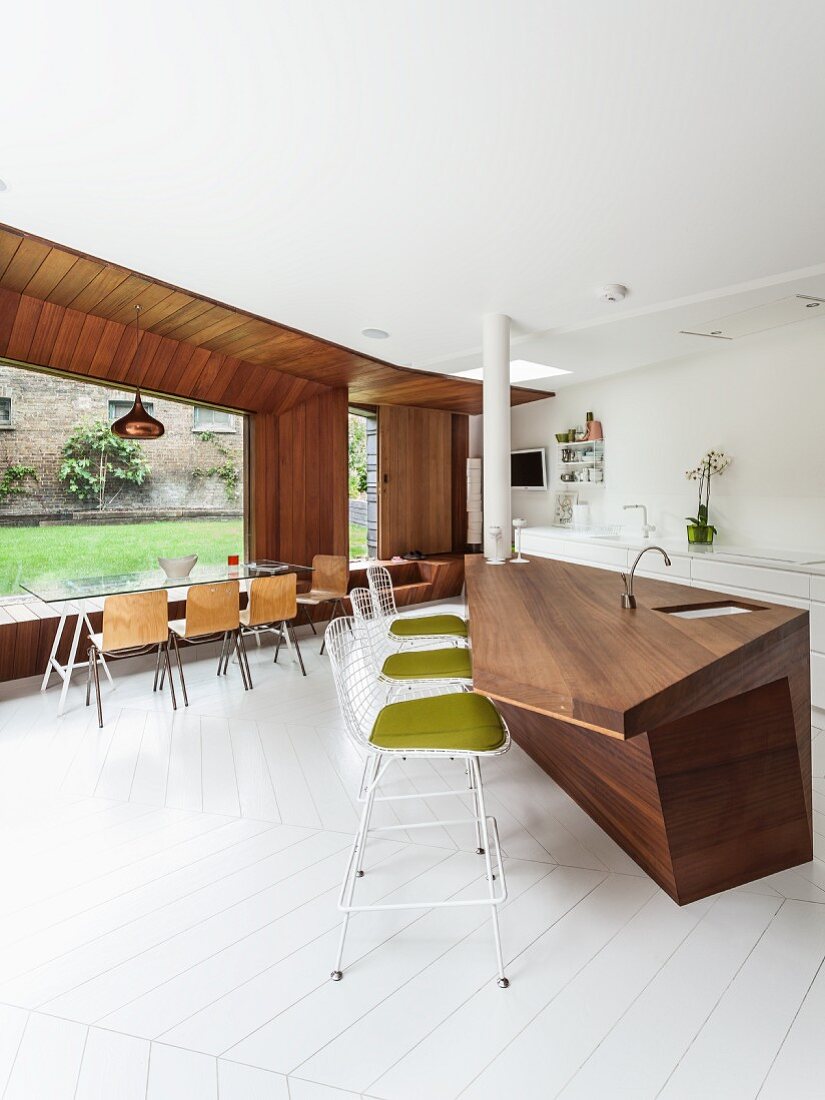 Küchentheke aus Edelholz mit weissen Barstühlen im Bauhausstil; im Hintergrund eine große Fensterfront mit langem Glastisch und hellen Holzstühlen