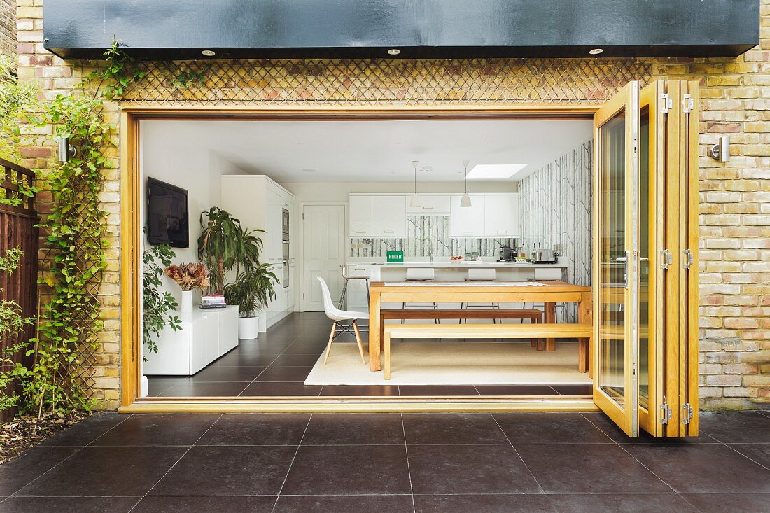 Blick durch offene Falttür in hellen Wohraum mit langem Holztisch und Sitzbank; im Hintergrund eine weiße Küchenzeile