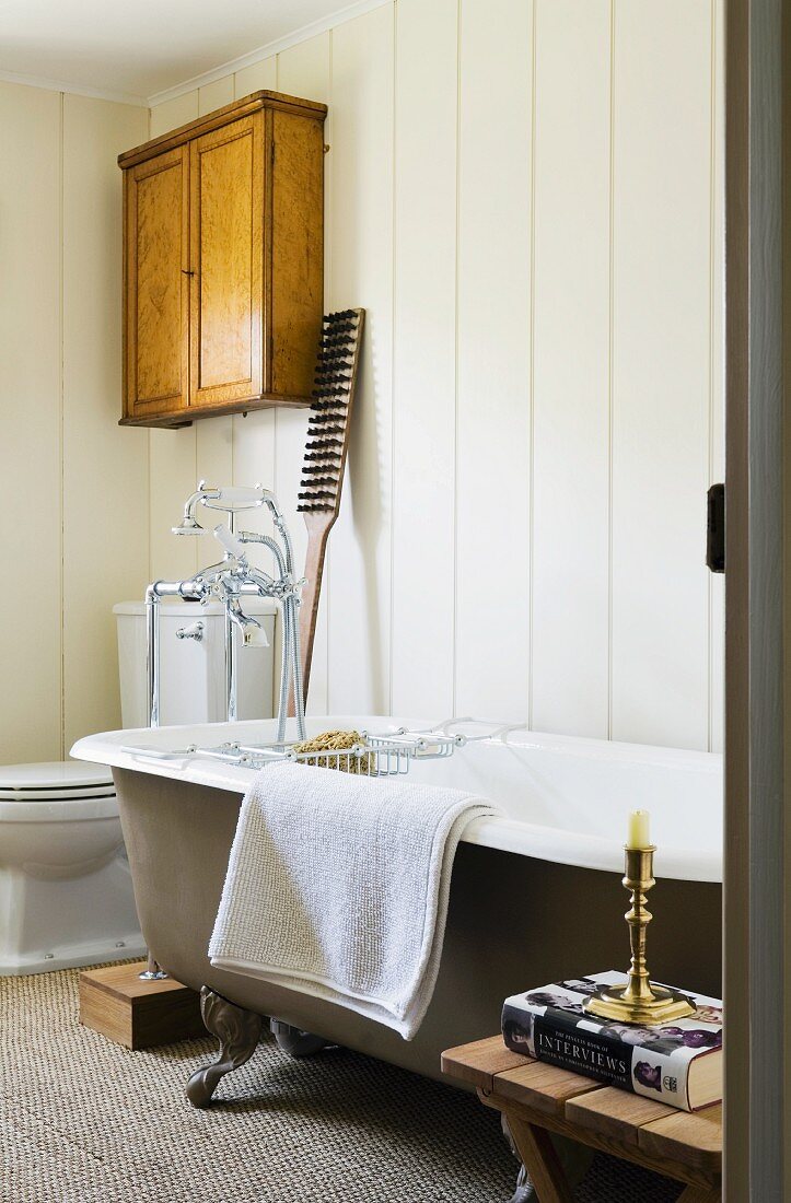 Freistehende Vintage-Badewanne vor holzverkleideter Wand, in der Ecke Toilette & antikes Wandschränkchen