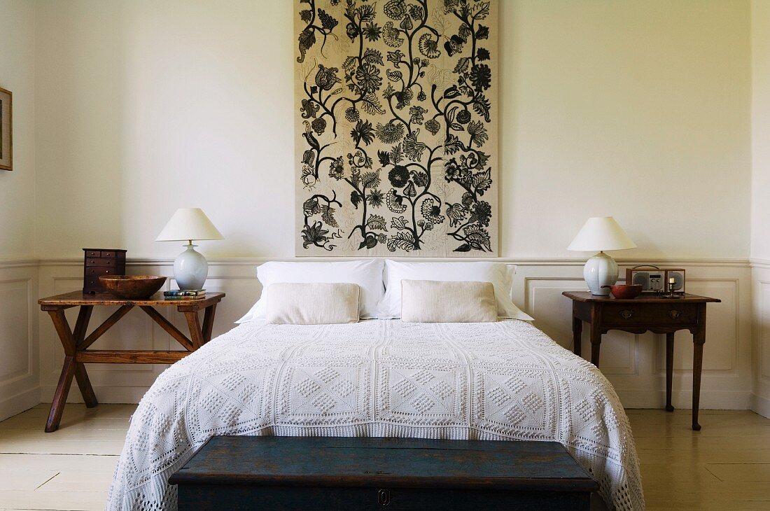Doppelbett mit Spitzen-Tagesdecke vor Wandpaneel mit Blumenmotiv, flankiert von Nachttischen in schlichtem Schlafzimmer