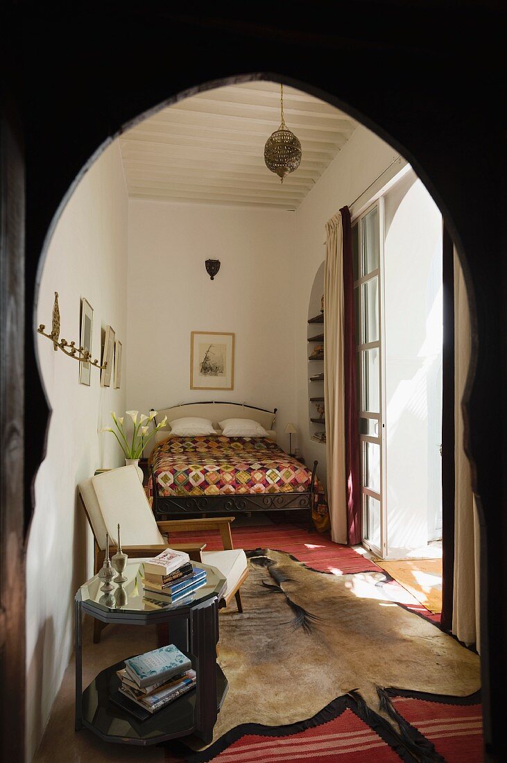 Blick durch orientalischen Durchgangsbogen in marokkanisches Schlafzimmer mit Bett, Sessel, Beistelltisch und Tierfellteppich