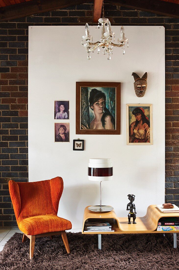 Fifties-style, orange armchair next to table lamp on coffee table in front of pictures hung on white panel on brick wall