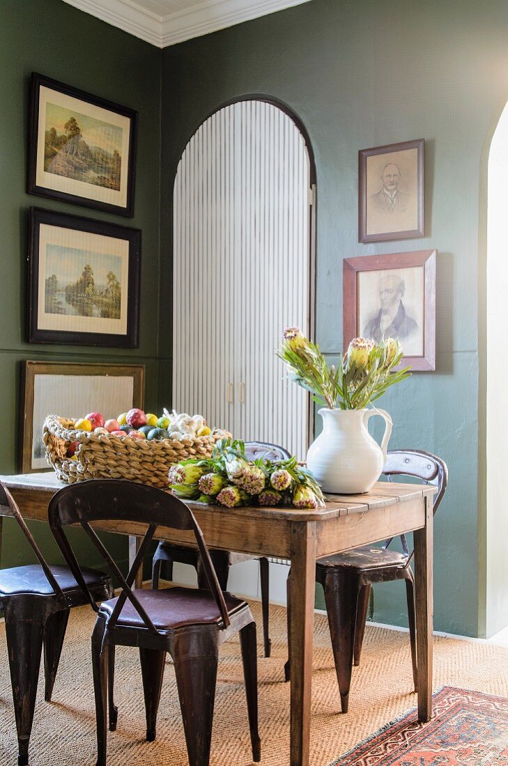 Flowers on rustic wooden table and retro-style metal chairs in corner of traditional dining room with framed pictures on green walls