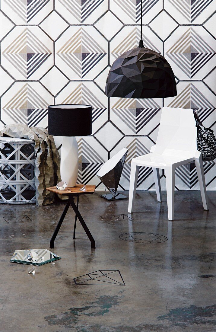 Black and white interior with geometric wallpaper behind white plastic chair and black pendant lamp; table lamp on three-legged side table in foreground