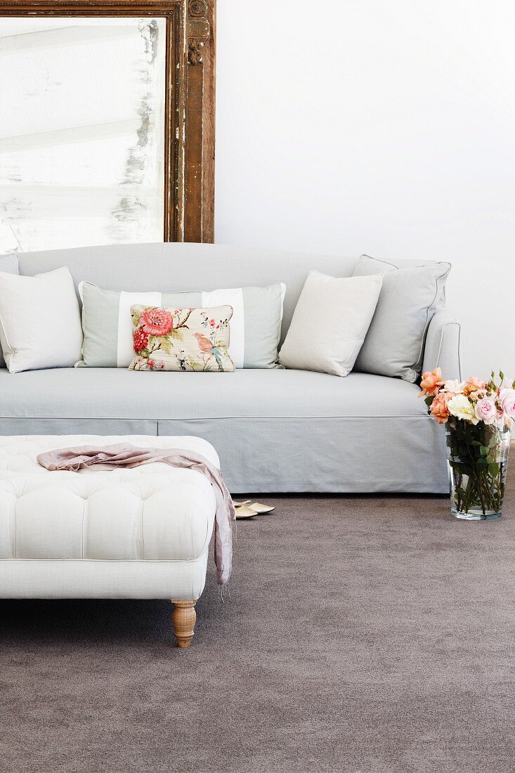Glass vase of roses on grey velour carpet next to elegant, pale sofa with scatter cushions and ottoman with wooden legs in foreground