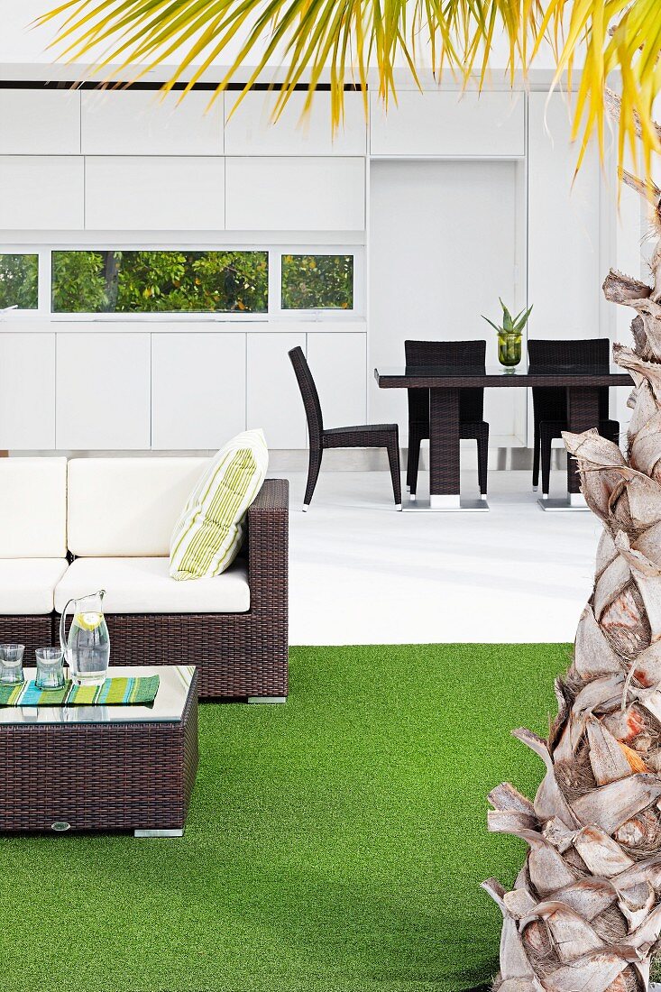 Blick über den Terrassenbereich mit Loungemöbel, Kunstrasen und Palmenstamm zum Eßbereich vor weißer Einbauküche mit Fensterband ins Grüne