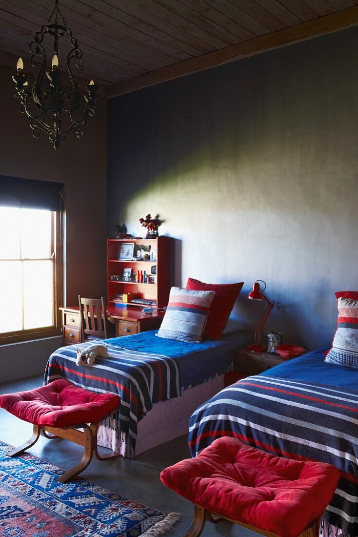 Colourful, striped bedspreads on twin beds and stools with red cushions in simple room