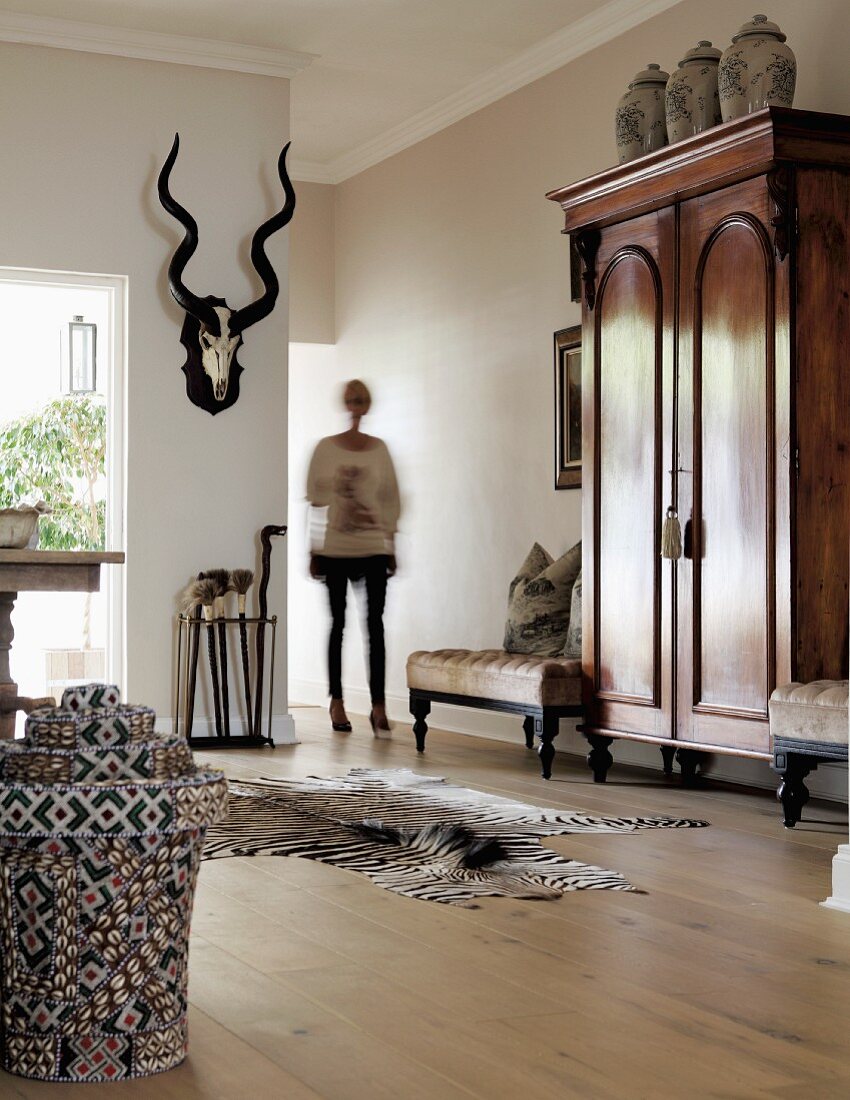 Elegantes Wohnzimmer mit antikem Schrank & buntem Rattankorb vor Zebrafell auf Dielenboden, im Hintergrund Frau neben Tiertrophäe an Wand