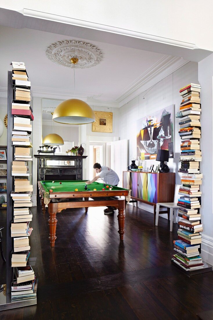 View of man playing billiards through wide open doorway flanked by stacks of books