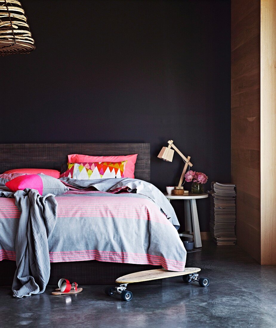 Skateboard on floor in front of double bed with headboard against black wall