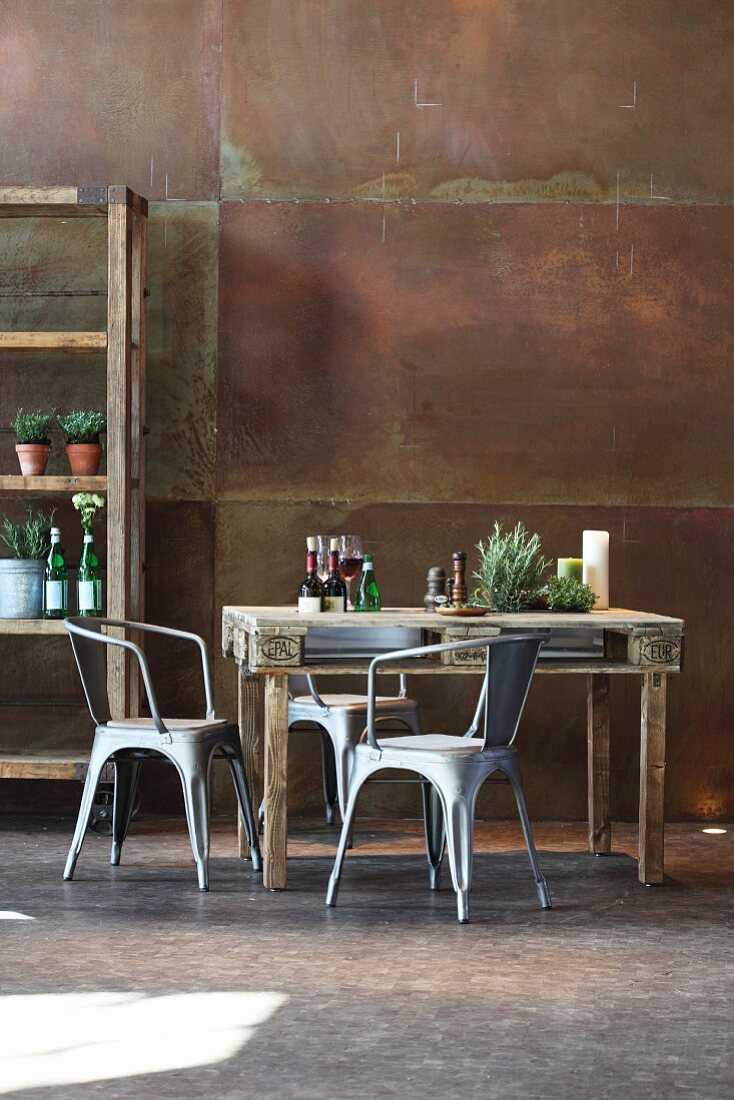 Rustic dining table made from pallet, metal chairs and partially visible shelving against corten steel wall
