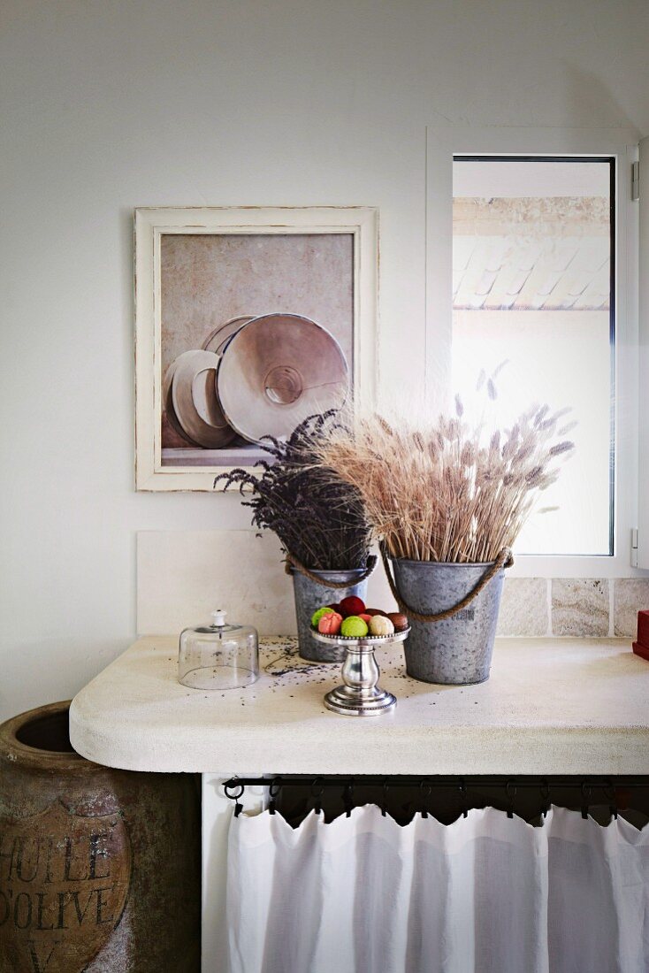 Dried flowers in metal buckets on worksurface with rounded corners below window