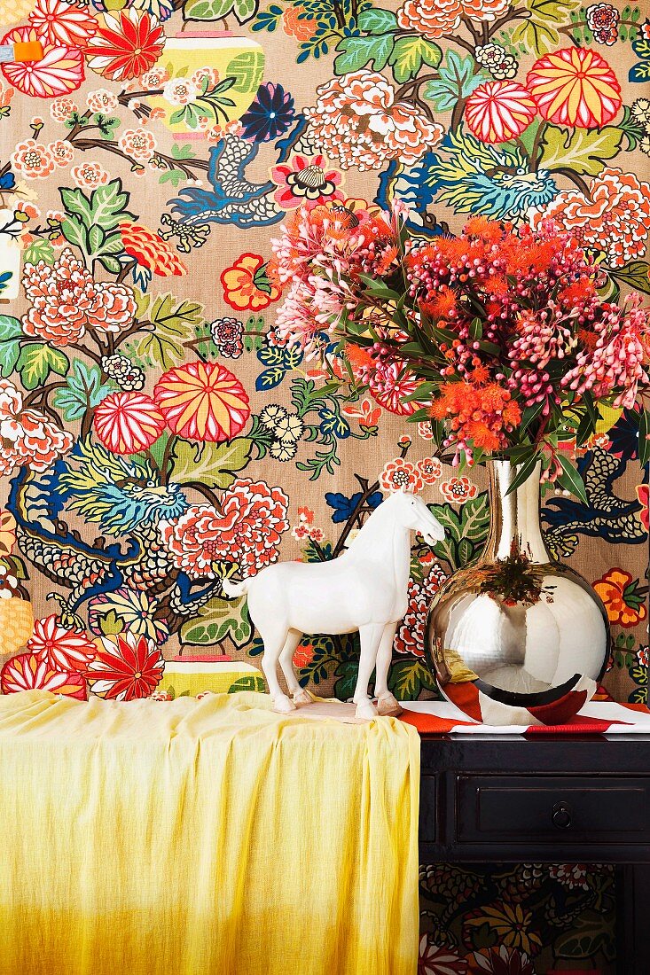 weiße Pferdefigur neben verchromter Vase mit Blumenstrauss vor Tapete mit farbenfrohem Muster