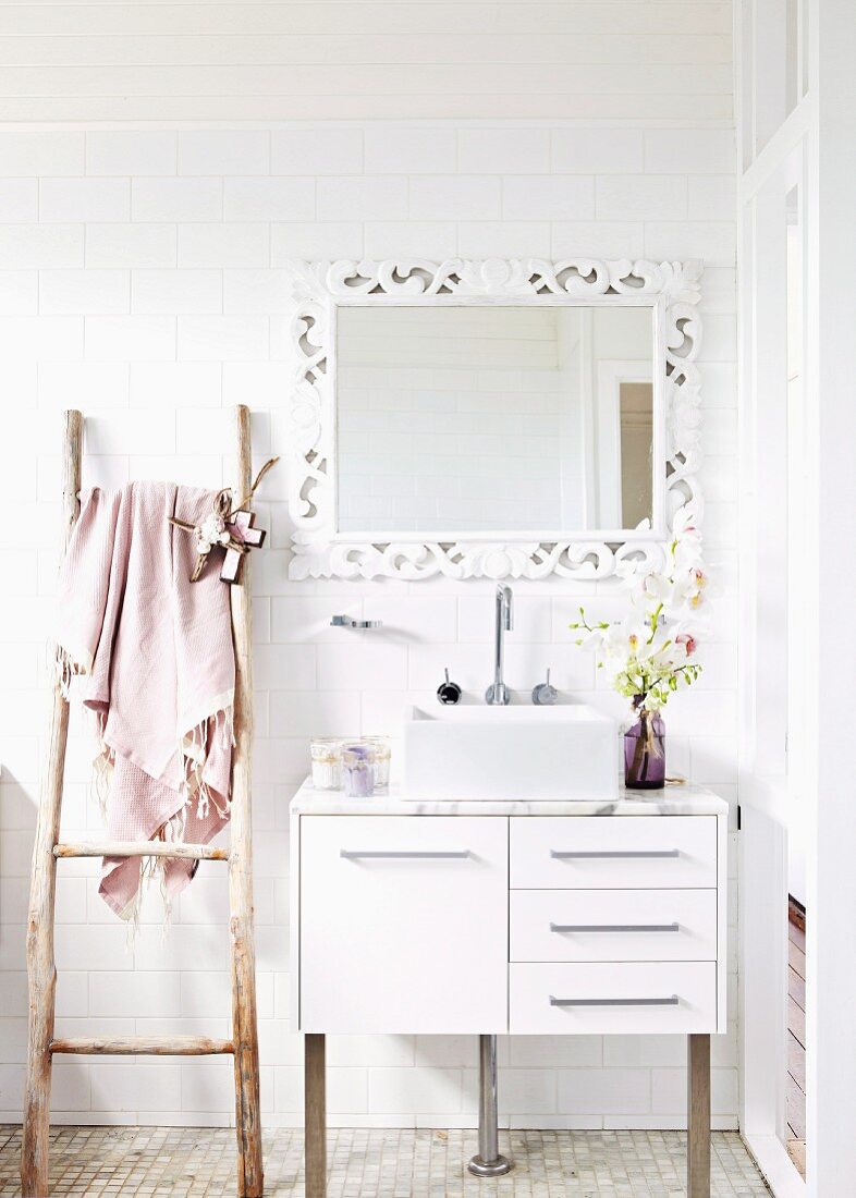 Waschbecken mit Schrankunterbau vor weissen Wandfliesen und gerahmtem Spiegel, daneben rustikale Holzleiter als Handtuchhalter