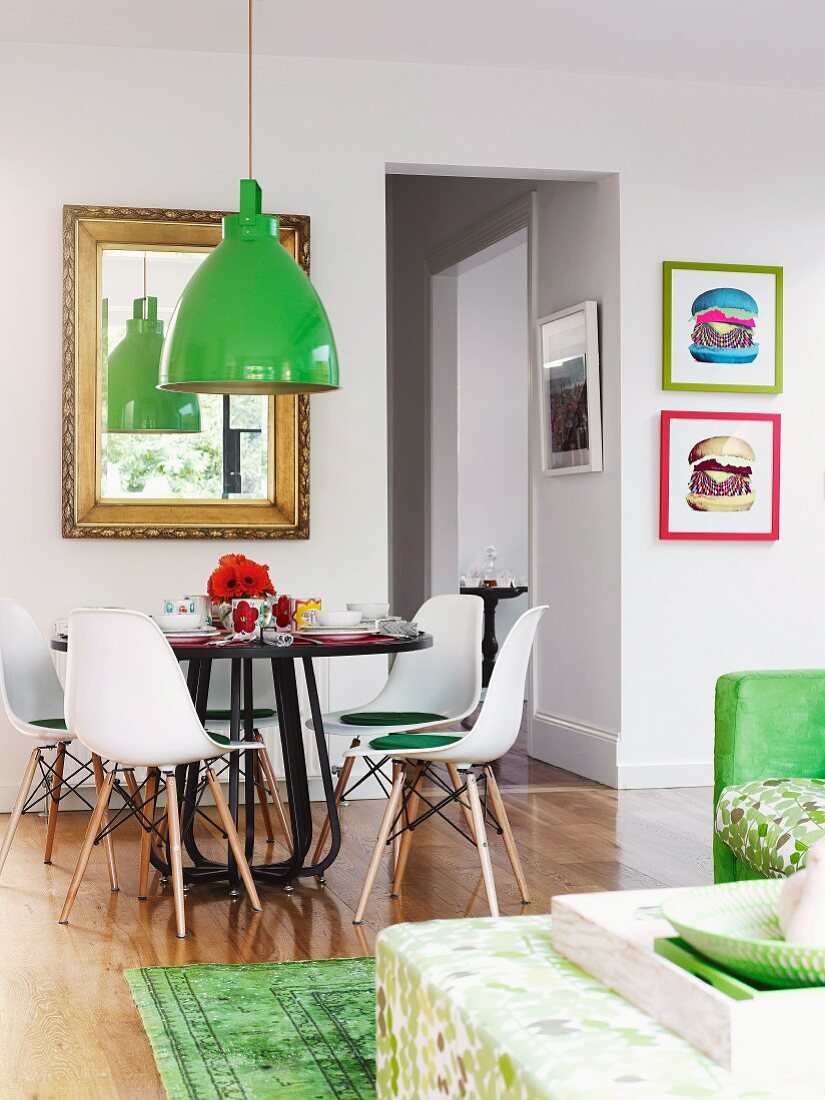 Grüne Pendelleuchte über Esstisch mit Klassikerstühlen; goldgerahmter Spiegel und bunte Burger-Bilder an den Wänden