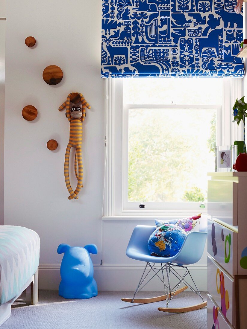 Blaue Akzente im Kinderzimmer mit gemustertem Faltrollo, Hundekopf und Klassiker-Schaukelstuhl; geringelter Spielaffe an der Wand