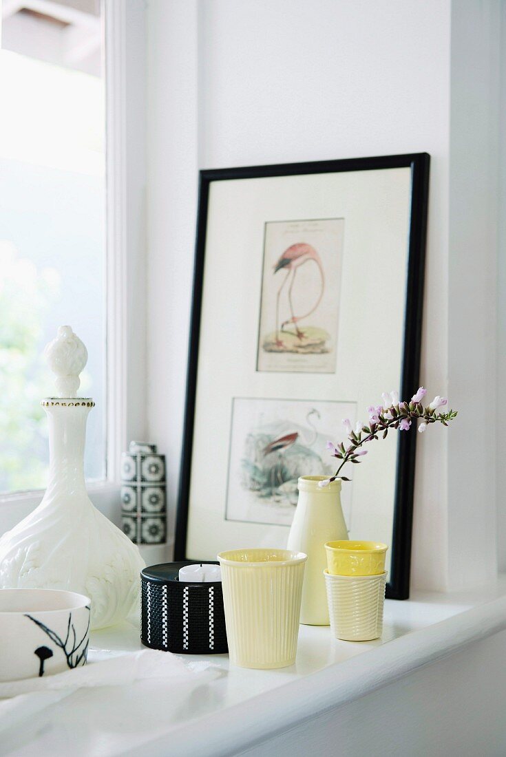 Verschiedene Becher aus Keramik und Vase mit Blumenzweig neben gerahmten Zeichnungen auf Fensterbank
