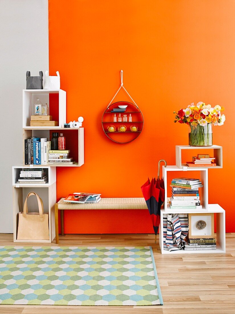 Stacked, cubic shelving modules & round ornamental shelving on wall above cloakroom bench against orange wall