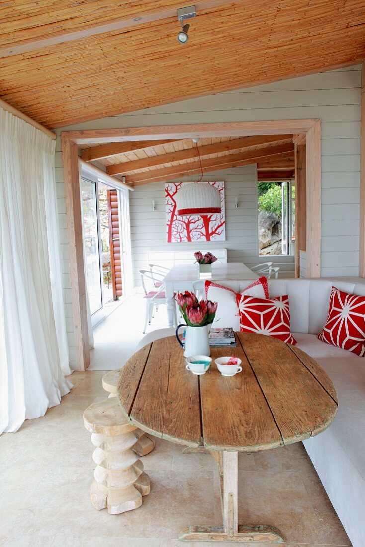 Rustikaler Holztisch und weisses Sofa auf überdachter Terrasse mit weissen, bodenlangen Vorhängen; Blick durch offene Tür ins Esszimmer