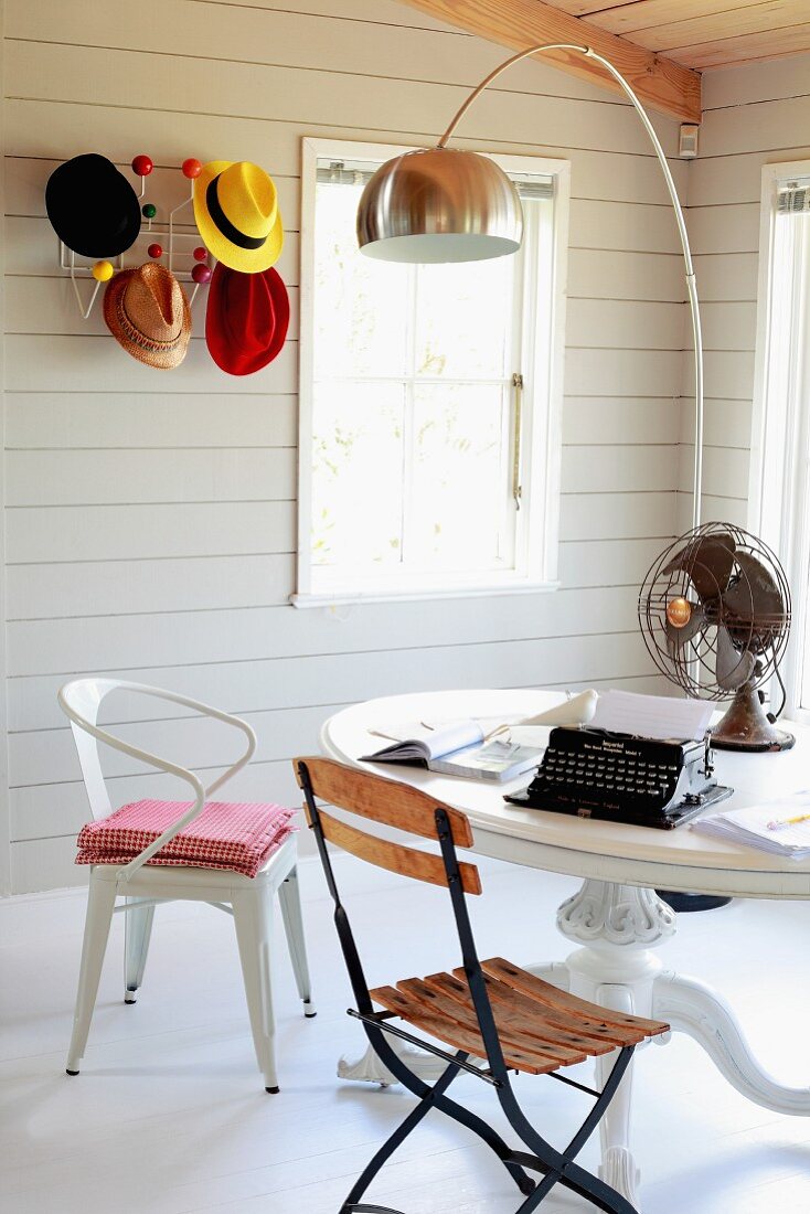 Verschiedene Stühle vor rundem Tisch unter Bogenlampe; an Wand Bauhaus Garderobe mit farbigen Hüten