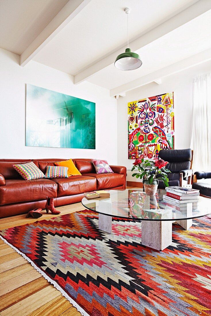 Runder Glas-Couchtisch auf folkloristisch gemustertem Teppich vor brauner Ledercouch in Wohnzimmer mit weisser Holzbalkendecke