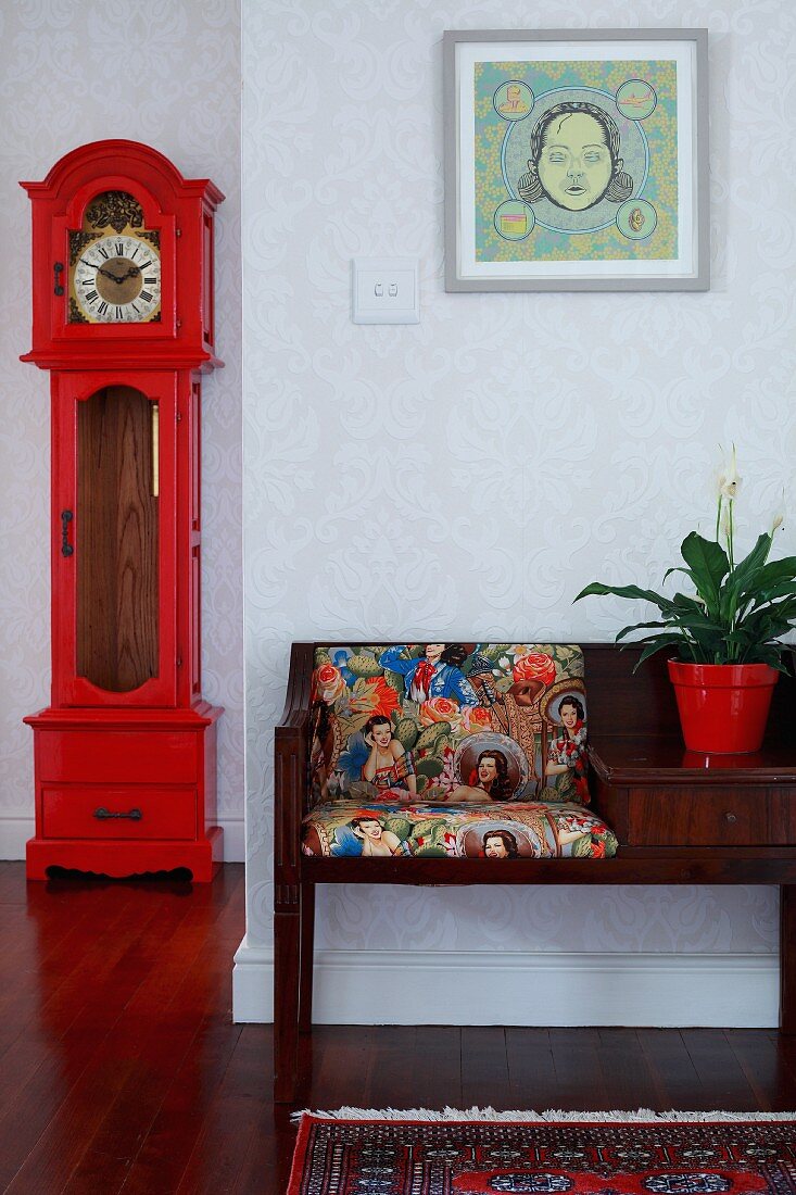 Telefonbank mit Musterkissen, gerahmtes Bild und rot lackierte, antike Standuhr in tapezierter Diele