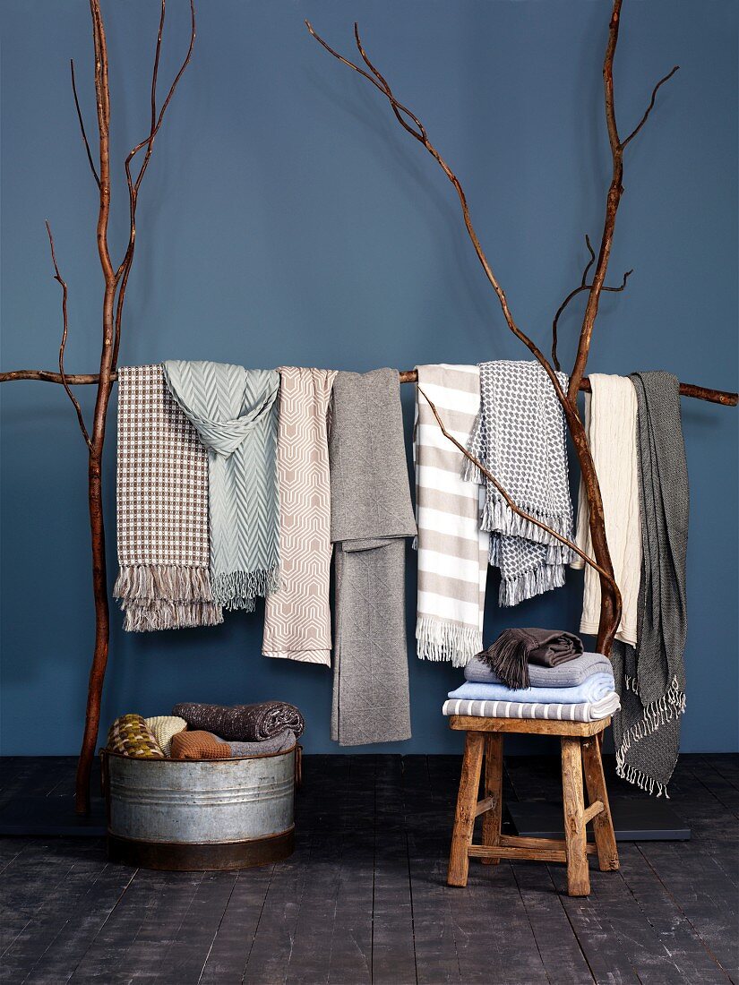 Schals und Decken in hellen Naturtönen vor blauer Wand - auf Gestell aus langen Ästen drapiert, in Zinkwanne und auf Hocker gestapelt