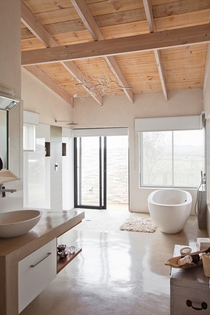 Weisses, modernes Bad mit Holzdecke und freistehender Wanne vor Fenster, daneben offene Regendusche mit Glasschiebetür ins Freie