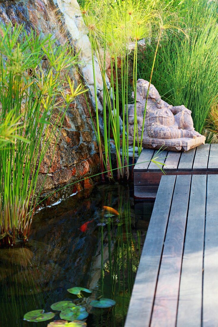 Steinfigur am Ende eines Holzsteges über Teich mit Wasserpflanzen