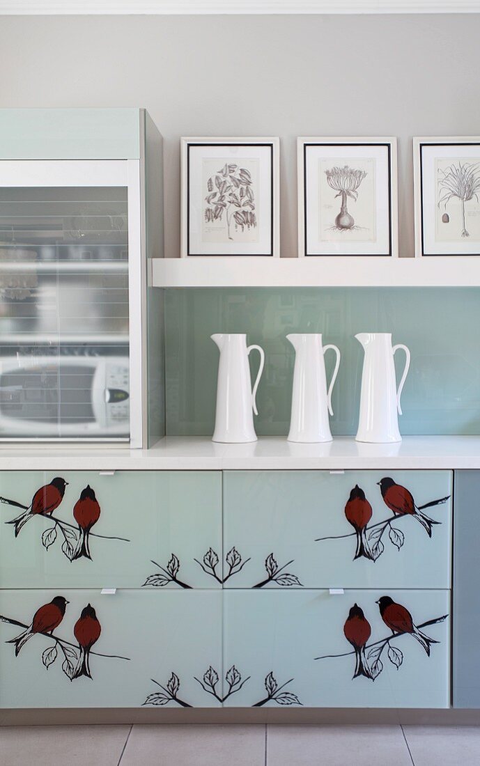 Schwarze Schattenriss-Vögel auf den Schubladenfronten eines Küchenunterschranks; weiße Porzellankannen und gerahmte Zeichnungen auf Wandbord