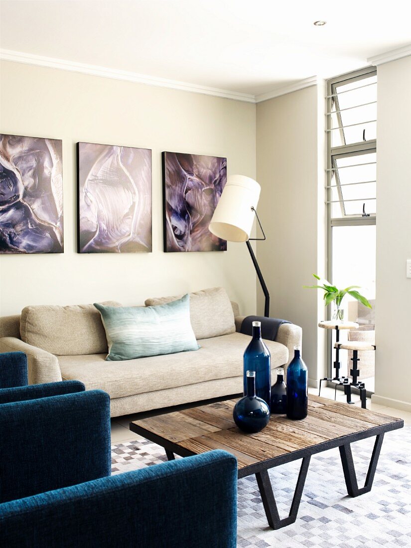 Corner of living room with organic photo prints above beige sofa and blue glass bottles on designer, wood and metal table