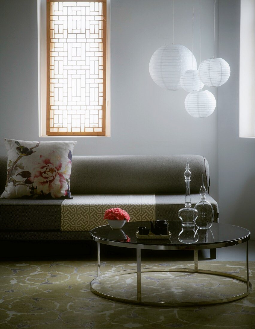 Retro-style, round side table in front of grey sofa and windows with Oriental-style screens flanking spherical pendant lamps