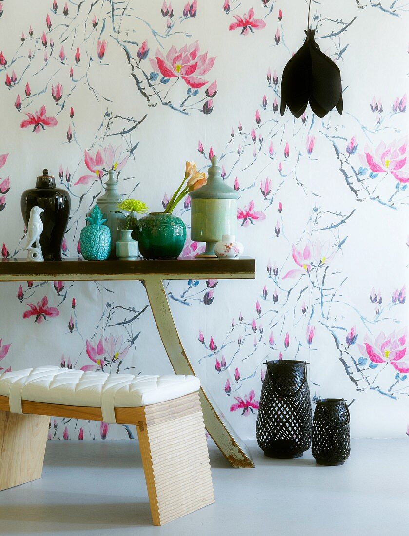 Gepolsterter Hocker und teilweise sichtbarer Tisch mit verschiedenen Porzellanbehältern vor Wand mit stilisiertem Blumenmuster auf Tapete