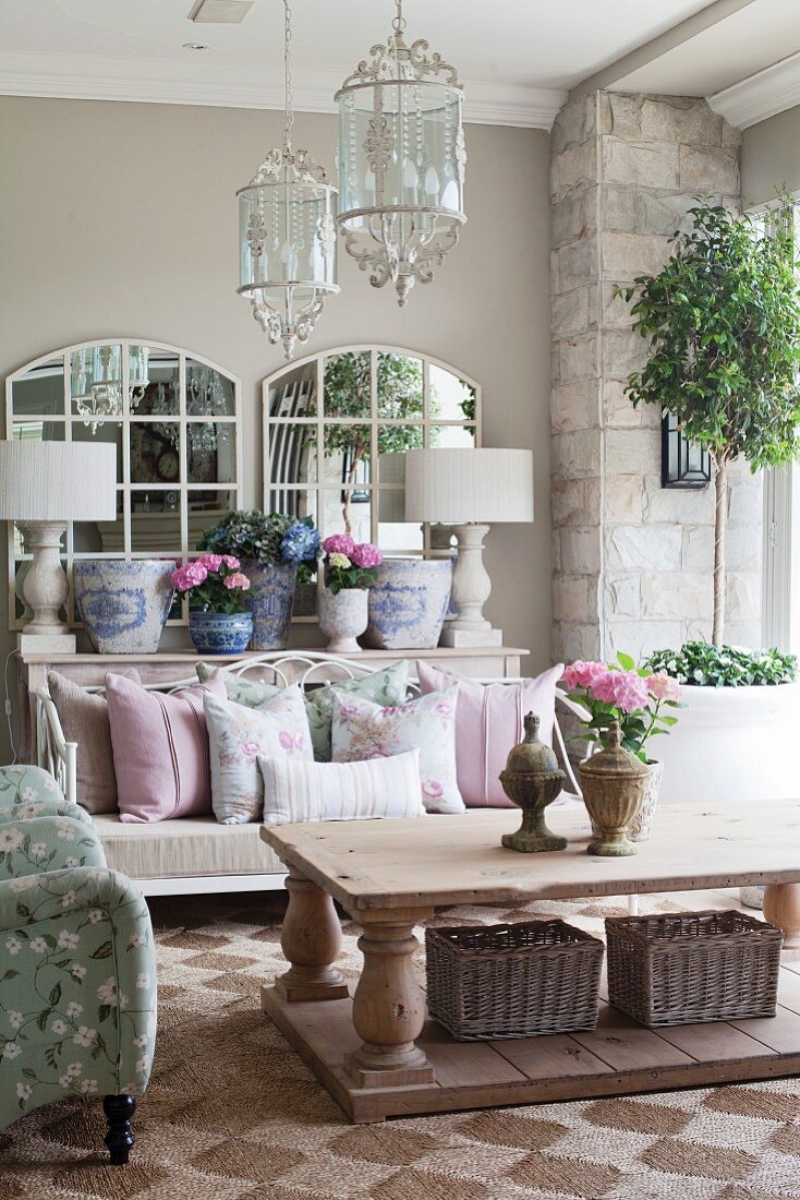 Eleganter Stilmix mit Metallbank und romantischen Kissen zu rustikalem Holztisch; zentrale Glasleuchter und Keramiktöpfe vor Sprossenspiegeln im Hintergrund