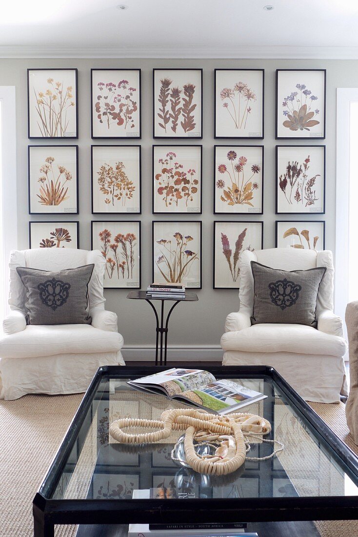 Couchtisch mit Glasplatte und weiße Polstersessel vor gerahmter Bildersammlung mit Blumenmotiven an Wand