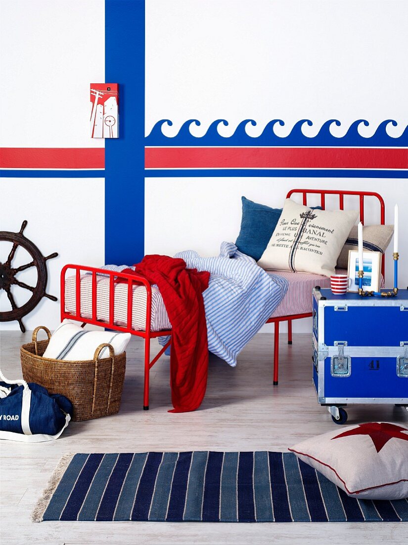 Vintage Bett mit rotem Metallgestell und mobiler Metallkasten in Blau mit Kerzenständern vor Wand mit gemaltem Wellenmotiv