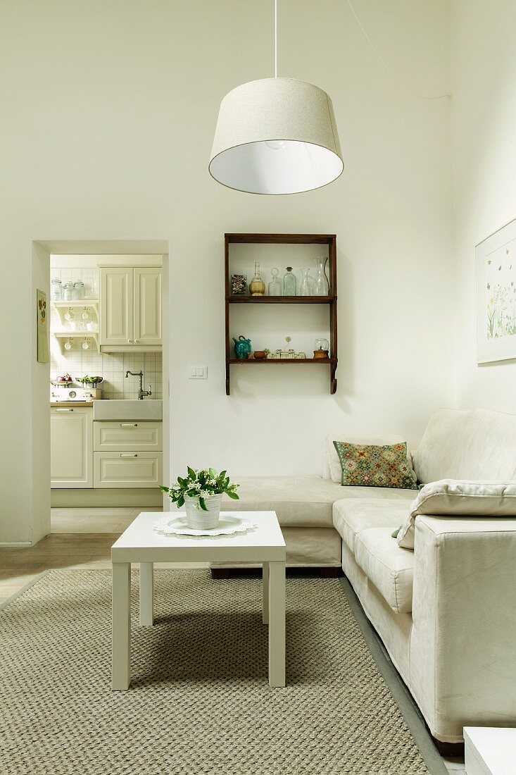 White coffee table on rug in front of pale sofa combination and simple pendant lamp in minimalist living room; doorway leading to kitchen in background
