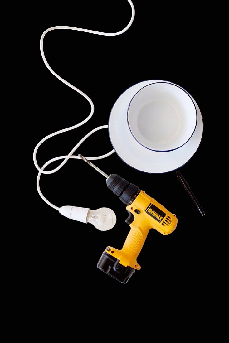 Gelber Akkubohrer mit Glühbirne, Stromkabel und weißer Emailschalen auf schwarzem Untergrund
