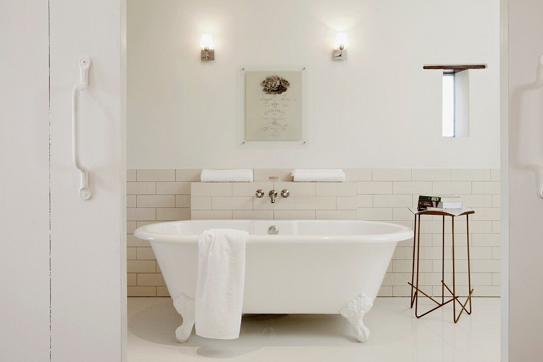 Blick durch offene, rustikale Schiebetür aus weißem Holz auf freistehende Badewanne mit Löwenfüssen und filigraner Metall Beistelltisch vor halbhoch gefliester Wand