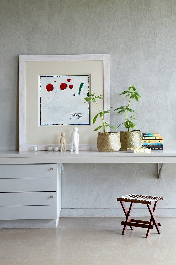 Modern artwork and houseplants in baskets on narrow console table with chest of drawers against grey marbles wall; stool with cord seat