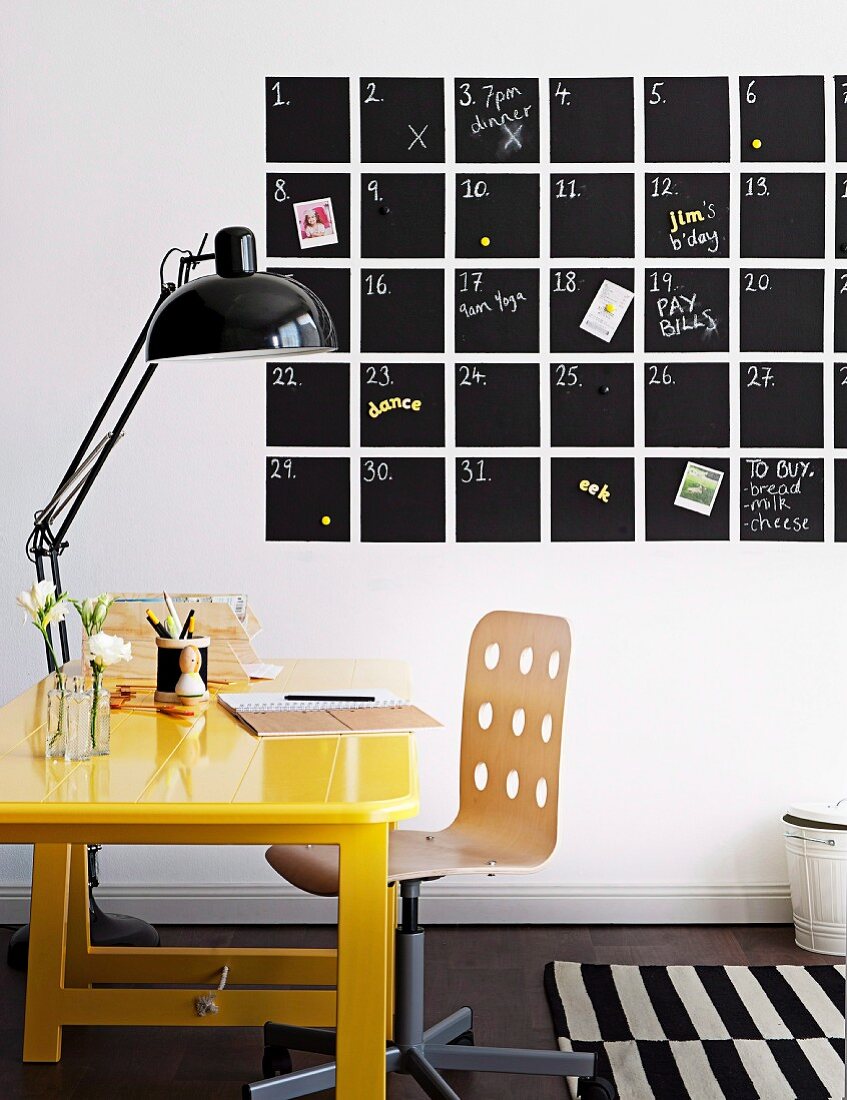 Gelb lackierter Schreibtisch und schwarze Retroleuchte vor Tafelkalender mit Eintragungen