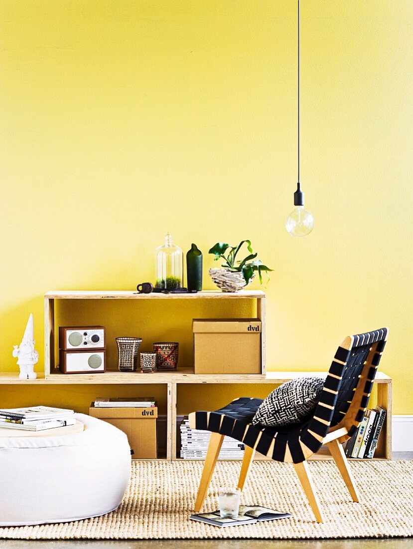 Selbstgemachtes Regal aus Sperrholz vor gelber Wand, im Vordergrund Retrosessel und Sitzkissen auf Sisalteppich