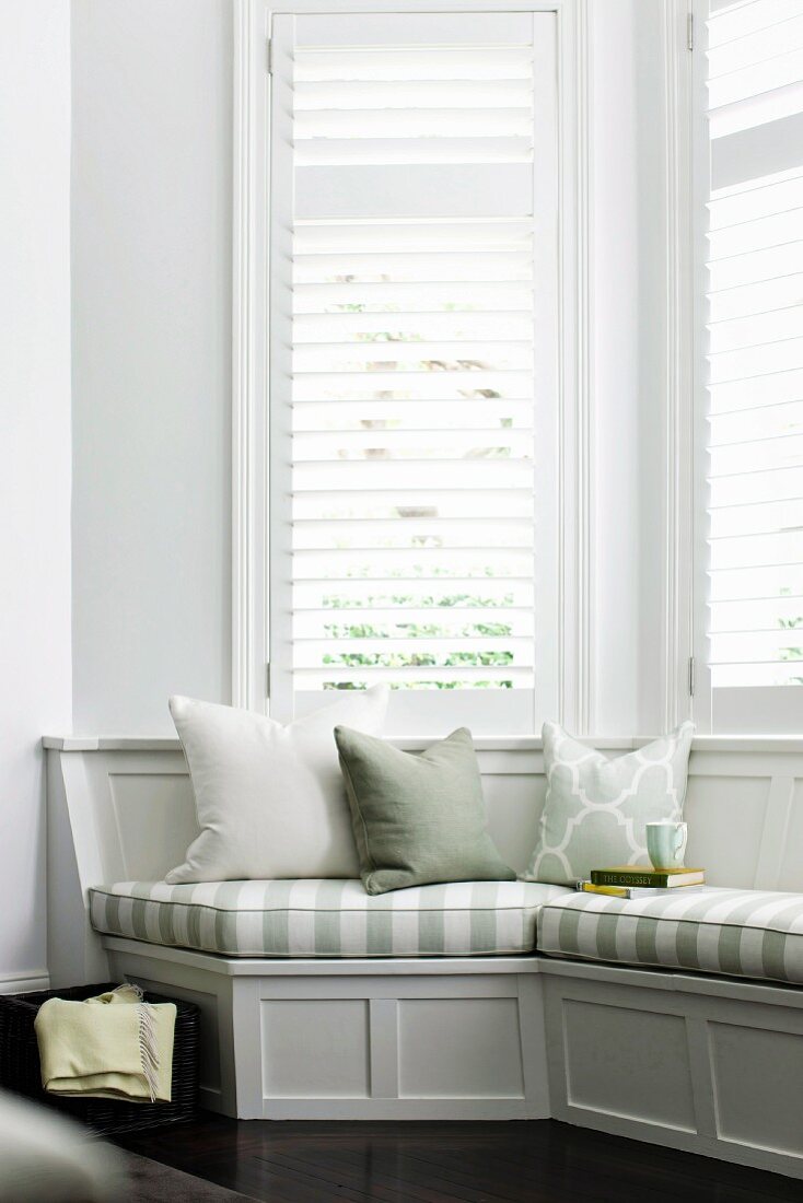 Gemütliche, elegante graue Holzsitzbank mit gestreiften Polsterauflagen vor weißen abgeschatteten Erkerfenstern läd zum Lesen und Verweilen ein
