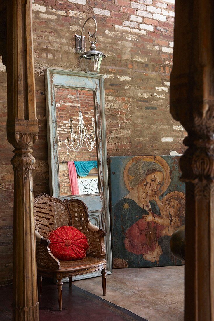 Antiker Geflechtsessel, Spiegel mit Vintage Holzrahmen und Mariengemälde vor rustikaler Ziegelwand, im Vordergrund geschnitzte Holzstützen