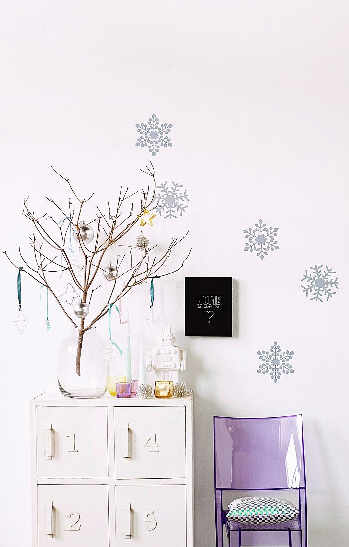 Weiß lackiertes Schränkchen mit winterlicher Dekoration an kahlem Ast in Glasvase, an der weißen Wand bläuliche Eiskristallmotive, darunter lilaner Plexiglasstuhl mit Kissen