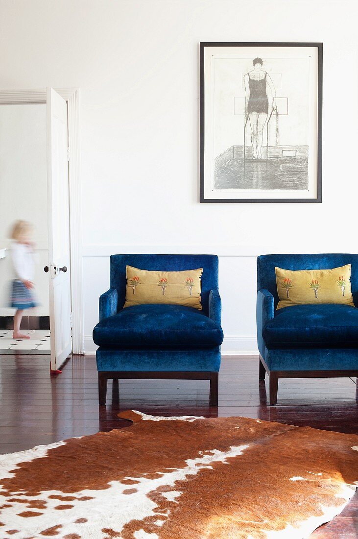 Tierfellteppich vor leuchtend blauen Sesseln und Kunstbild an der Wand; seitlich eine offene Tür