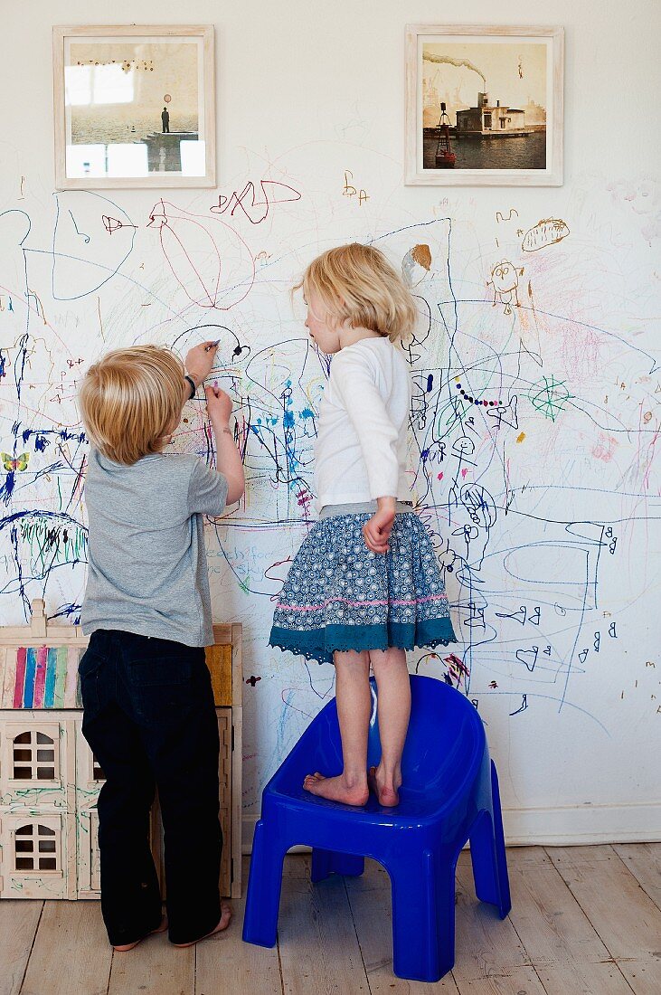 Zwei Kinder beim Bekritzeln einer weissen Wand unterhalb gerahmter Fotografien